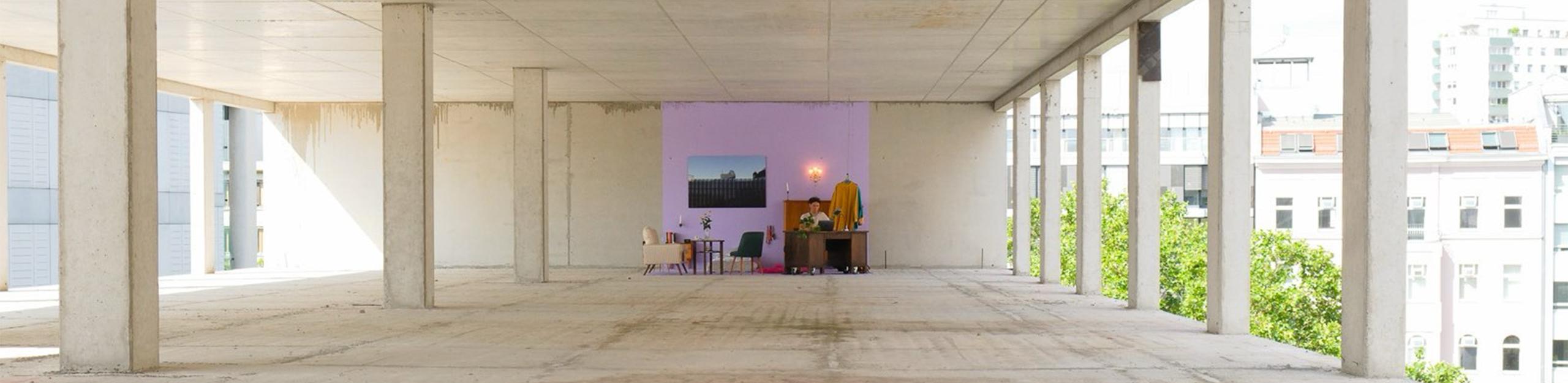 Zu sehen ist der Rohbau eines Bürokomplexes von SIGNA in Berlin, in dem Parasite Real Estate eine Intervention gestartet hat. Es wurde ein Teil der Wand und Boden in einem hellviolet gestrichen und Möbel a la einer Ein-Zimmer Wohnung auf die Fläche gestellt.